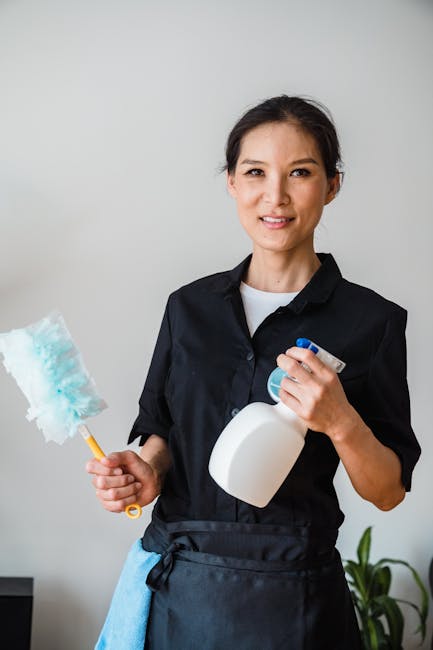A professional cleaner from hirecarpetcleaner.london, wearing a black uniform and black gloves, is wiping a wooden dining table with a light blue cloth in a well-lit, modern living space. The table su