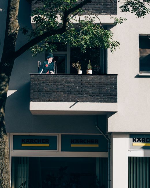 Image showing a residential building with a small balcony featuring a black brick railing. On the balcony, there are two potted plants, a person dressed as a chimney sweep in a traditional outfit, inc