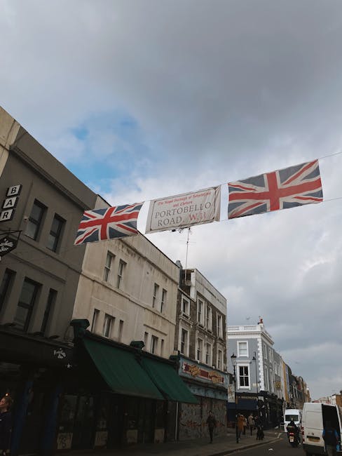 A street scene in Notting Hill showing a large banner hanging above the road, decorated with British Union Jack flags on either side. The banner reads 'Portobello Road Market Tips,' and the surroundin
